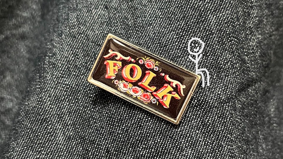 A stick figure sitting on a small enamel pin which says 'folk' on it